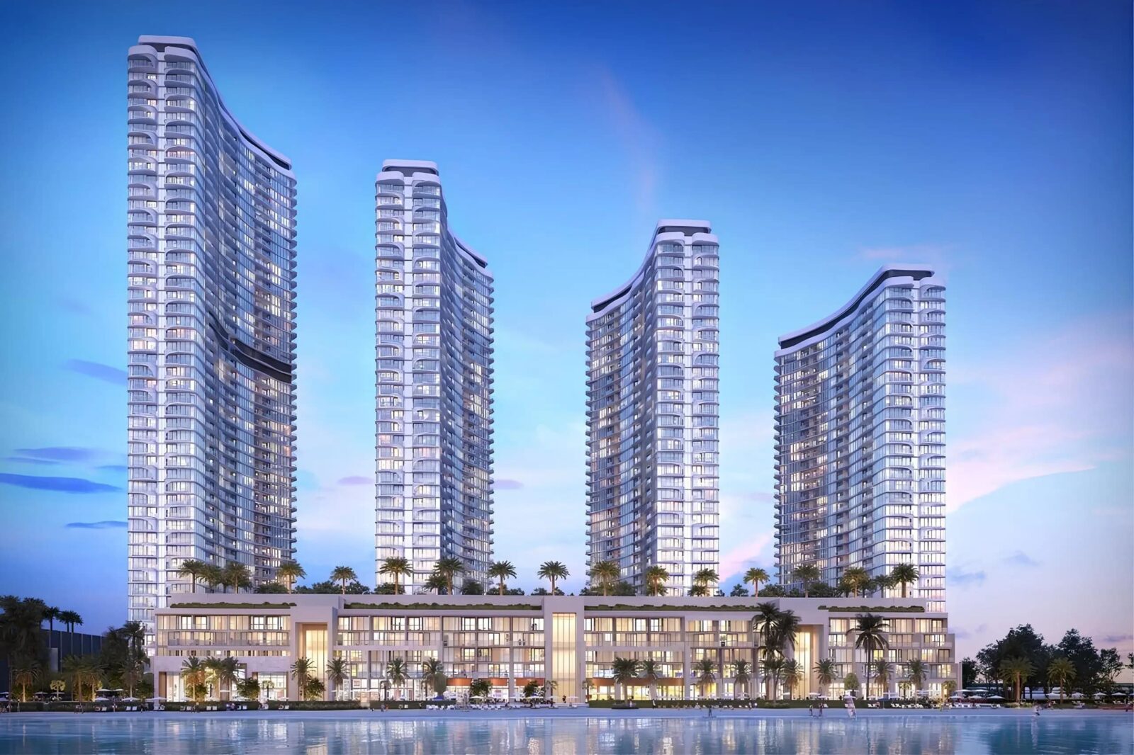 Evening view of the four illuminated Sobha Aquacrest towers with their wave-shaped roofline above the glass podium